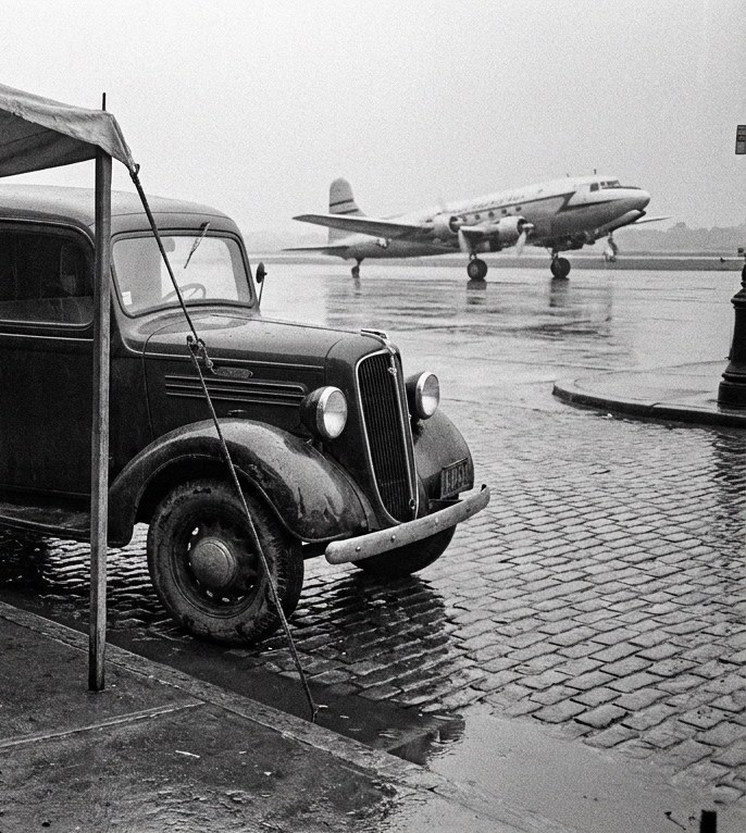 Black-and-white 1940s scene: a delivery truck on wet cobblestones with a propeller airliner on the airfield behind
